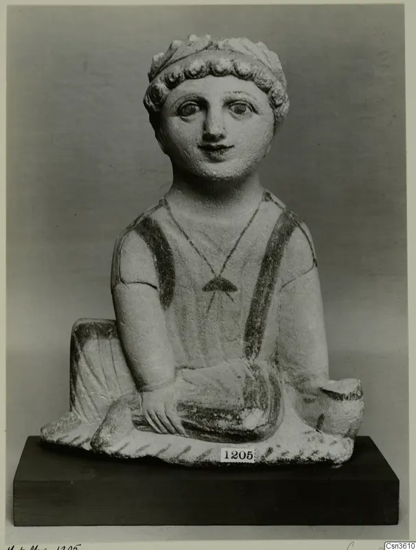 Seated limestone figurine with curly hair, painted eyes and dark painted straps, shown on a dark wooden base.