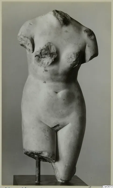 Marble torso of a standing human figure, headless and armless, mounted on a metal stand against a grey backdrop.