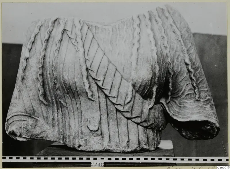 Marble sculpture fragment of a draped garment with vertical pleats and a diagonal band, shown on a dark stand.