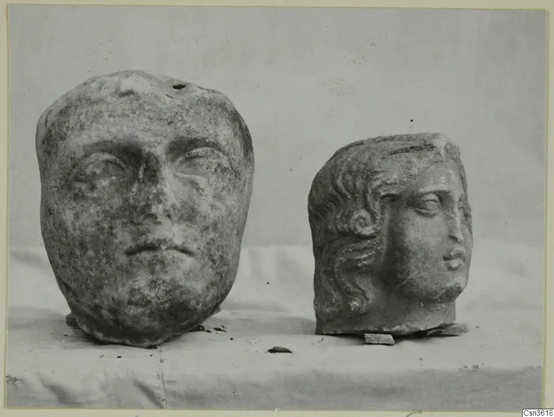 Two carved marble heads side by side on a pale cloth, one broad-faced and one smaller with curled hair