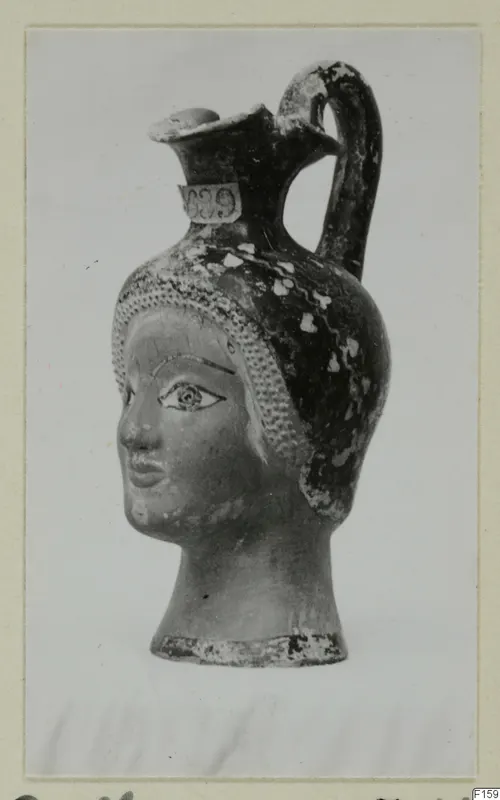 Jug shaped as a female head with a strap handle and trefoil mouth, shown on a mounted card