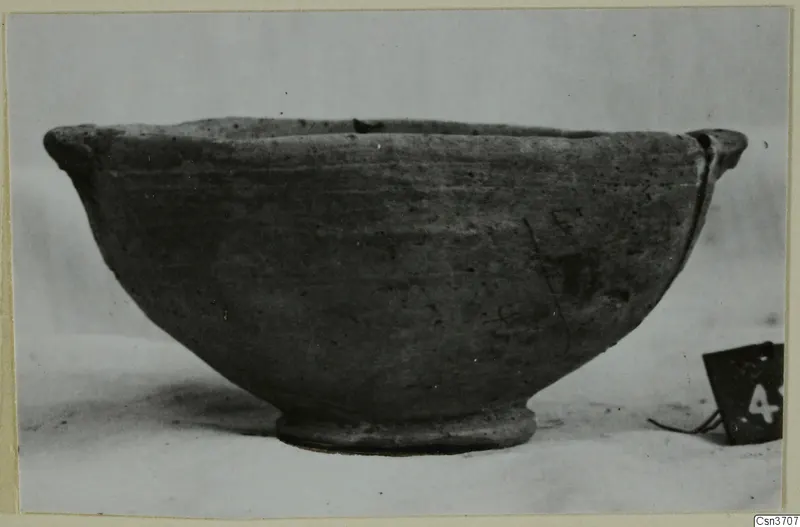 Grey polished ware bowl with a flaring rim, a small lug and a low ring foot, photographed from the side