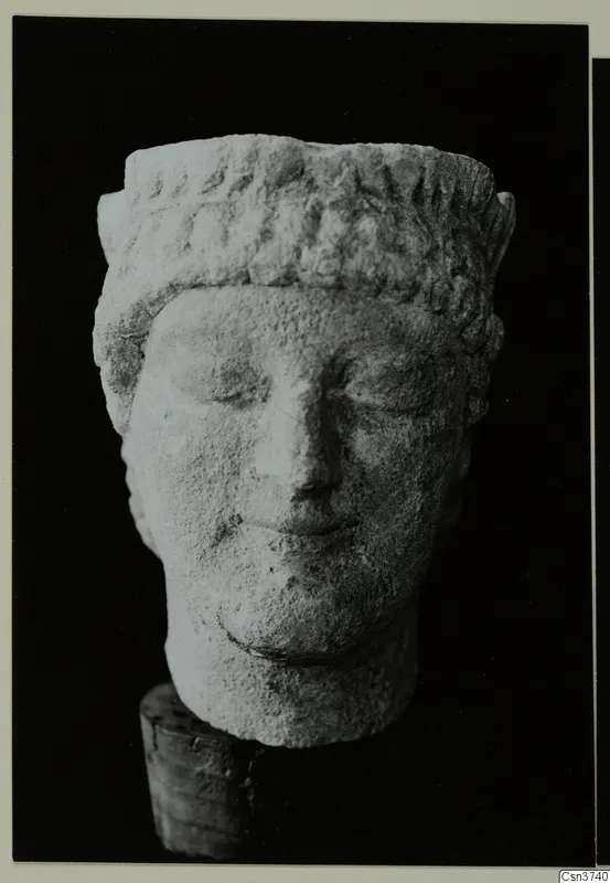 Limestone head of a sculpture from Mersinaki, with carved curls across the top and a worn face, set on a wooden support.