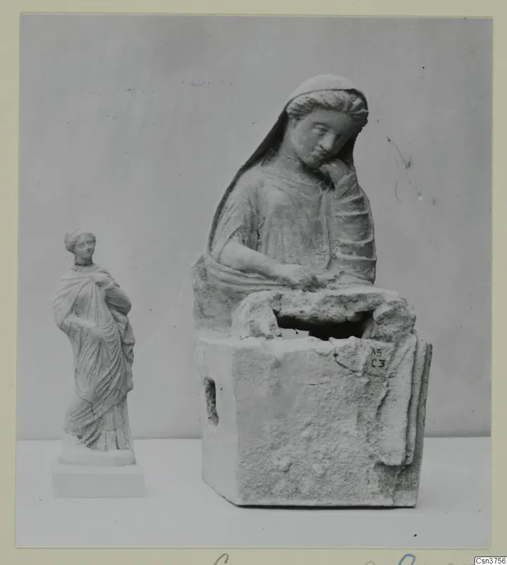 Two pale female figures photographed together: a larger seated figure on a block base and a small standing draped figure.