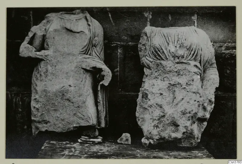 Two headless stone statues of draped standing figures against a dark wall, from Arsos