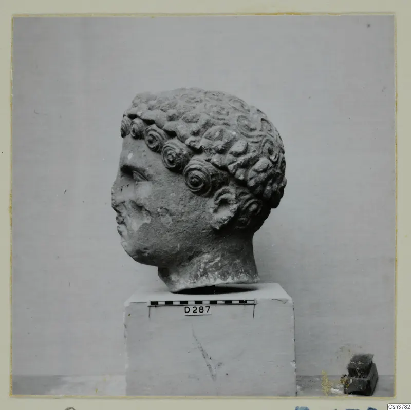 Roman sculpture: carved stone head with rows of curled hair, set on a block beside a scale bar