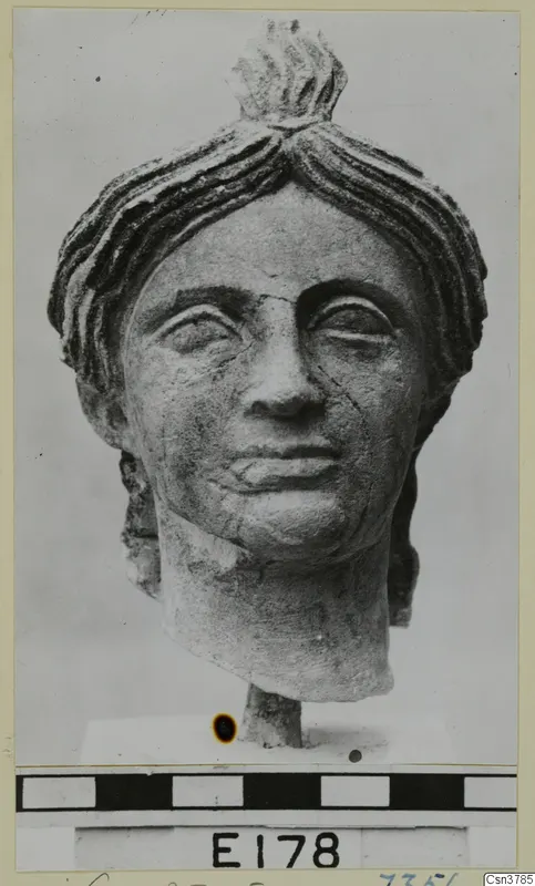 Carved stone head of a woman with hair swept up in a topknot, on a short base against a grey backdrop.