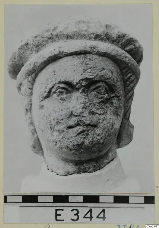Stone head sculpture seen from the front, with a thick rolled headdress, large eyes and a pitted, worn surface