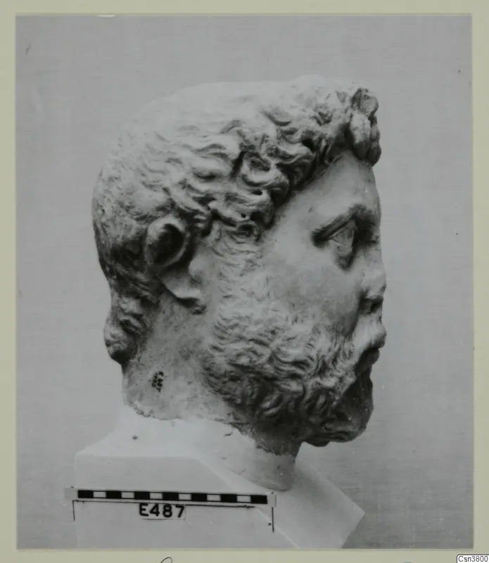 Head of a bearded, curly-haired man carved in stone, shown in right profile on a plinth with a scale bar