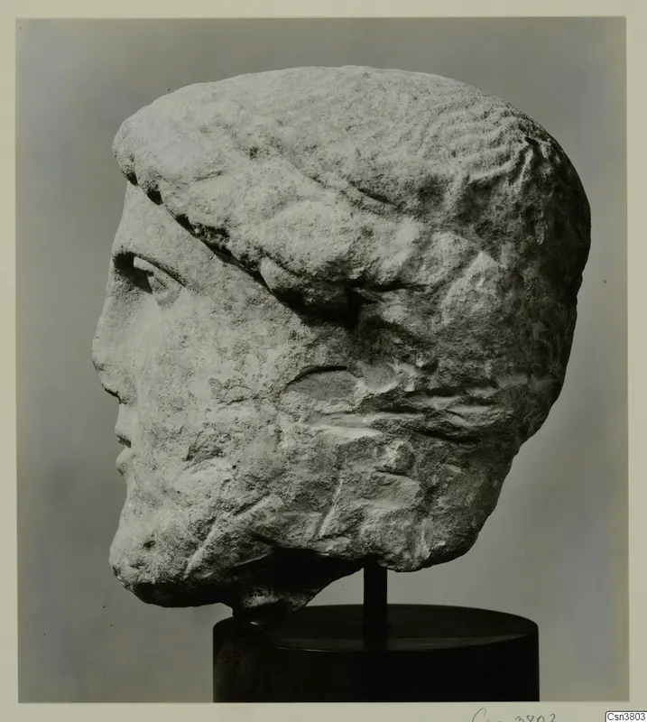 Carved stone male head in profile, with a band across the hair, mounted on a black stand.