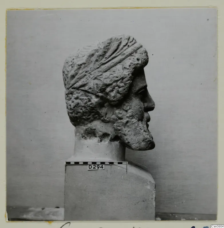 Carved stone head of a bearded man in profile on a square base, with a scale bar and number label.