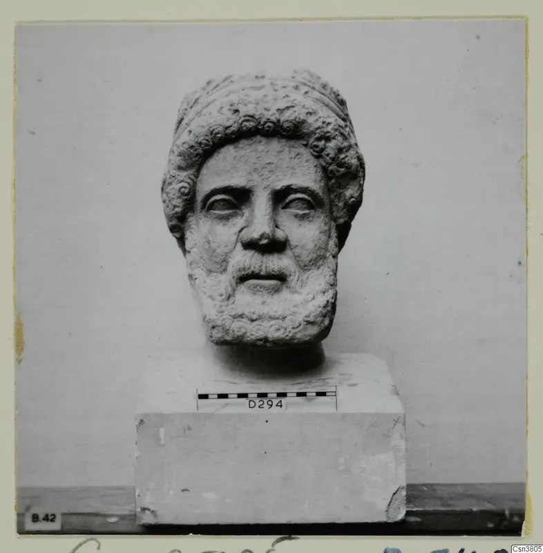 Carved stone head of a bearded man with curled hair and beard, set on a block base
