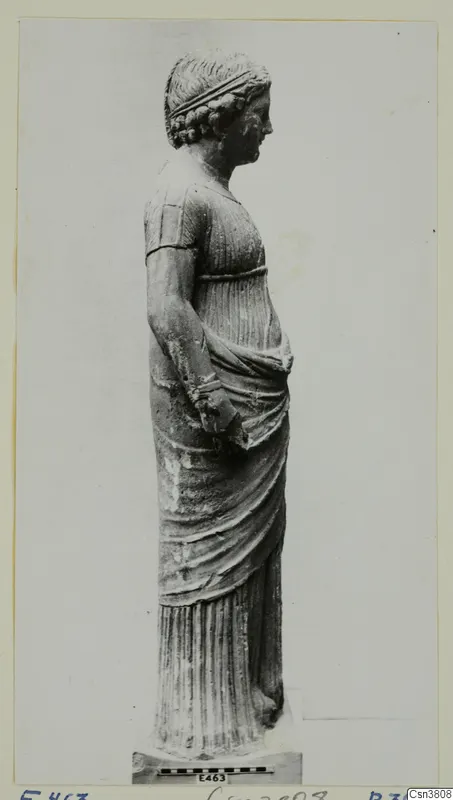 Standing stone statue of a draped female figure, shown in profile, with a measuring scale bar at its base.