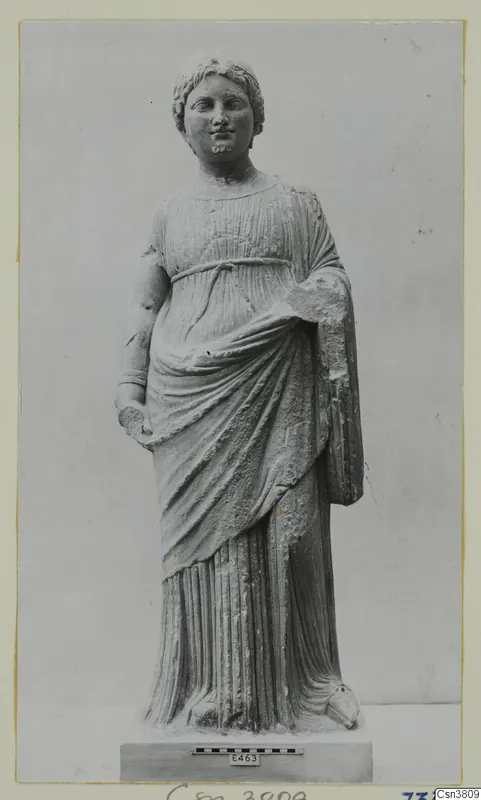 Stone statue of a standing female figure in a belted, draped dress, shown on a plinth with a scale bar.