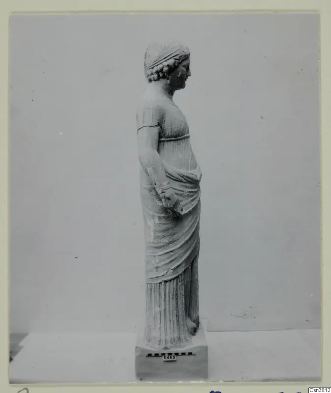 Standing female statue in stone, shown in profile facing right on a rectangular base.
