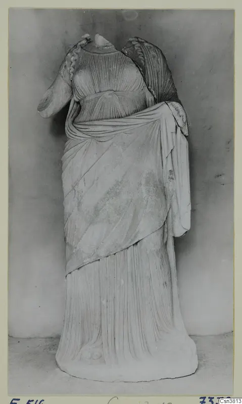 Standing draped statue in stone, head and neck missing, photographed against a grey backdrop