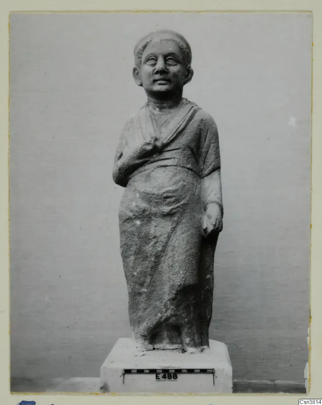 Standing stone statue of a draped figure on a white plinth, with a scale bar and number label in front.