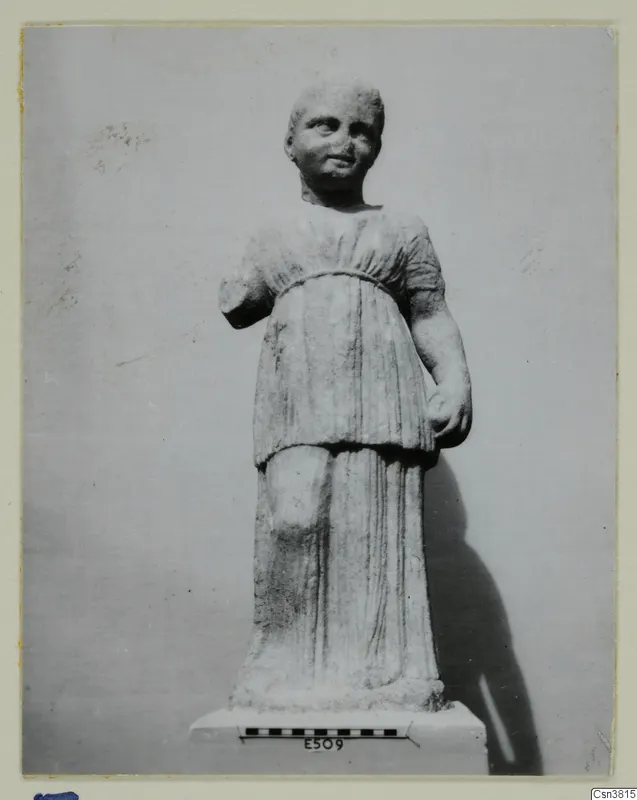 Carved stone statue of a standing figure in a belted, short-sleeved garment, on a base with a scale bar.