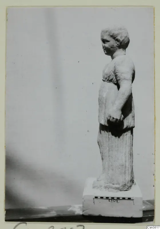 Pale stone statue of a standing figure in a belted tunic, shown in profile on a rectangular block base.