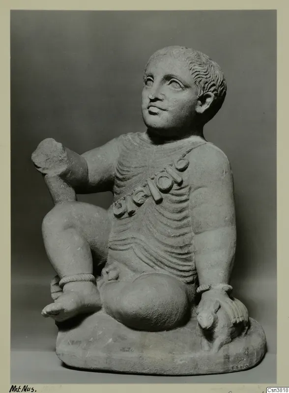 Stone statue of a seated child with a bead garland across the chest, photographed against a grey background.