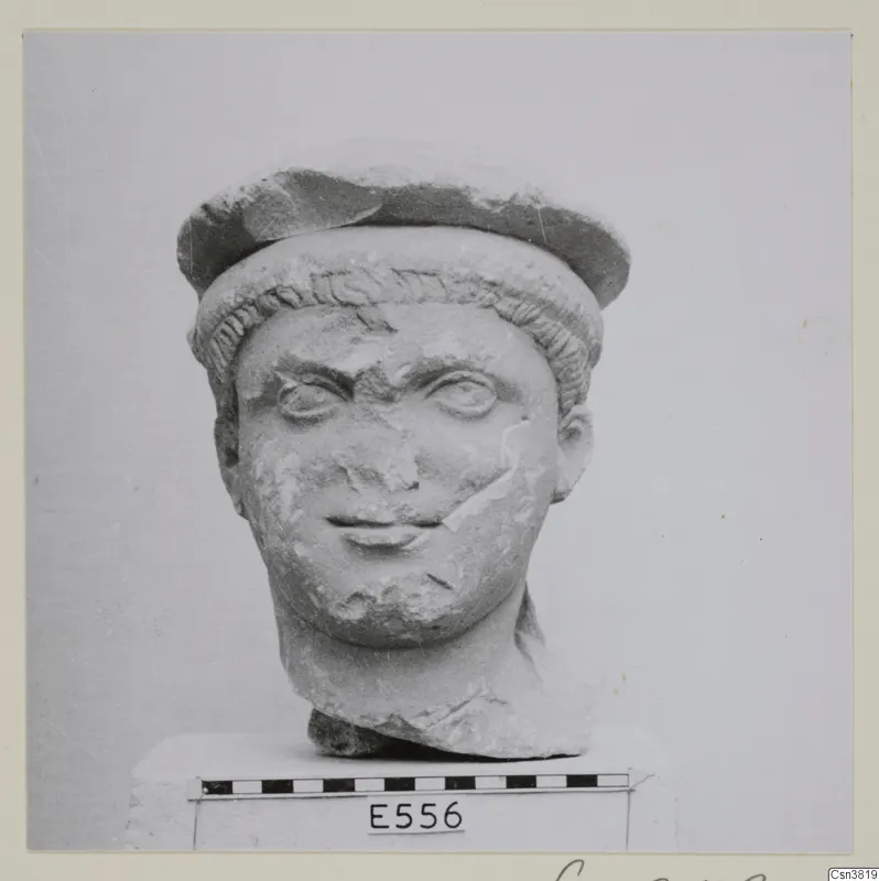 Carved stone head of a youth with a cap-like headdress, shown on a block against a plain background.