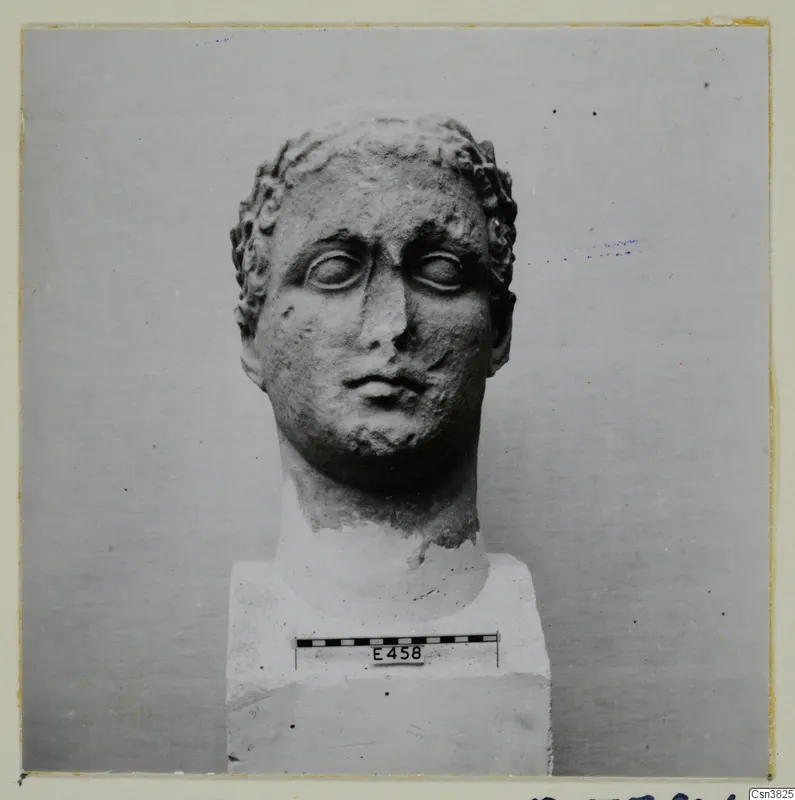 Carved stone head with curled hair, mounted on a square block, with a scale bar in front of the block.