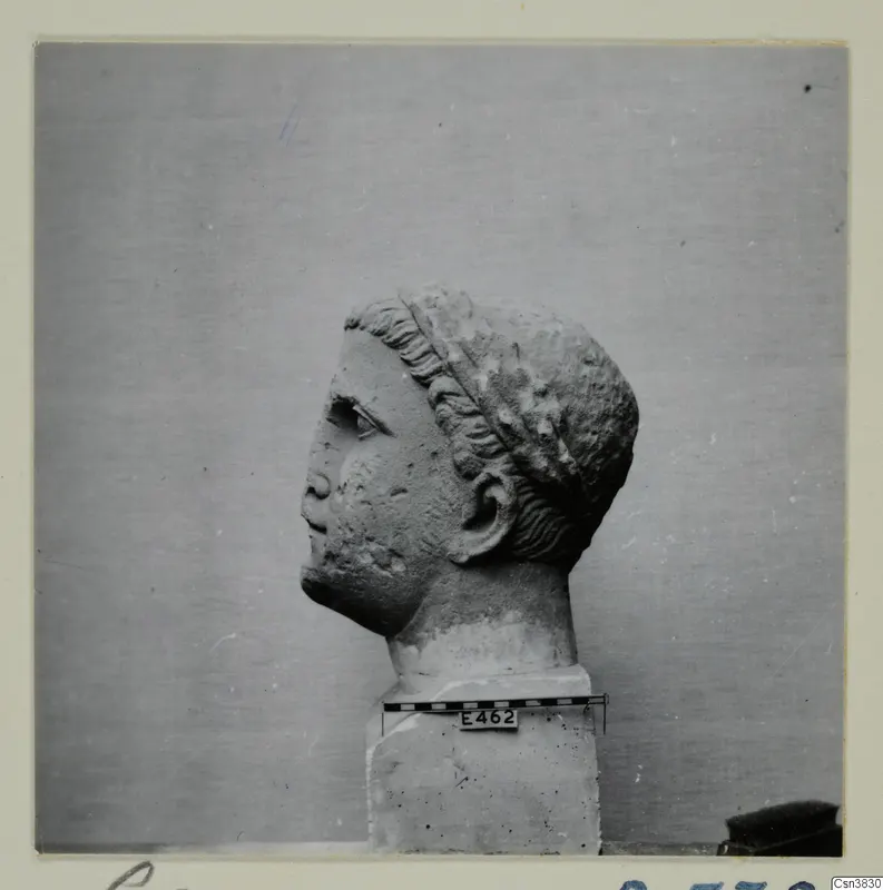 Stone head sculpture in profile, facing left, on a square base beside a scale bar.