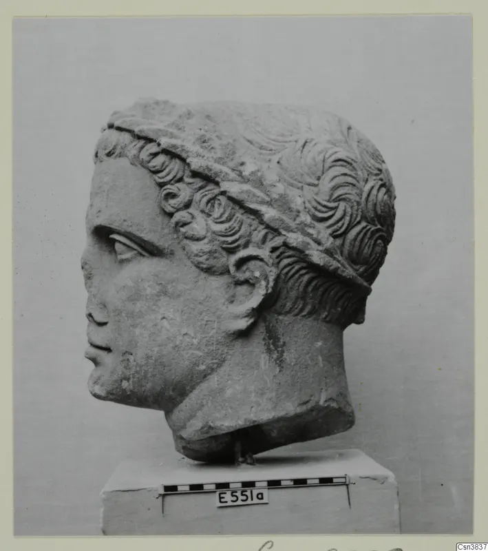 Carved stone head of a young figure in profile, with curly hair and a band across the head, on a plinth with a scale bar