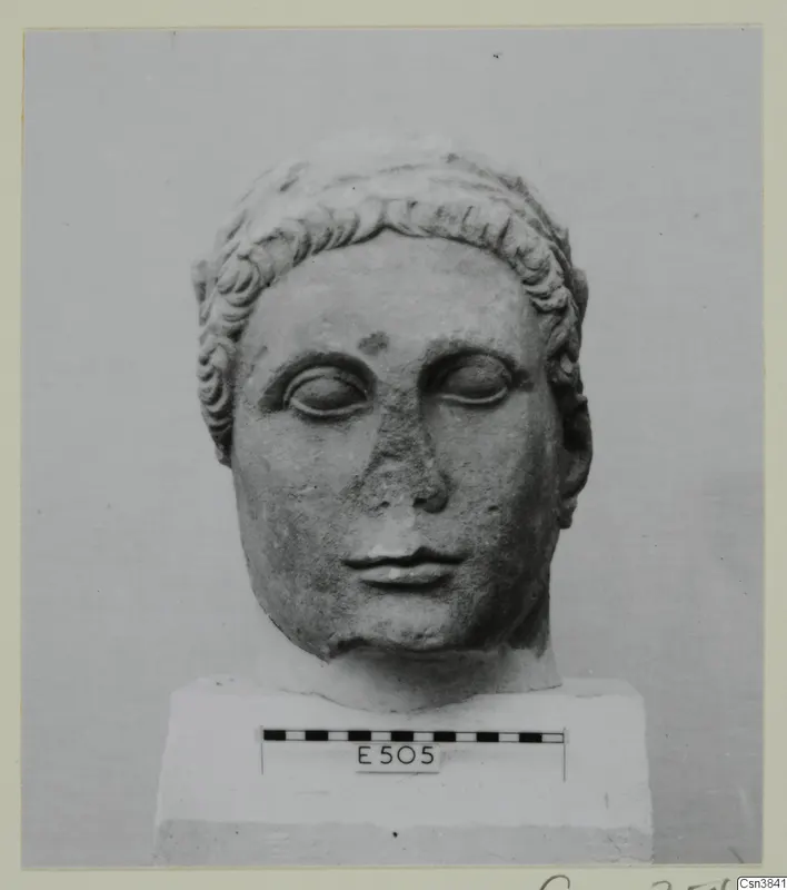 Carved stone head with curled hair and lowered eyes, standing on a plinth with a scale bar and number label.