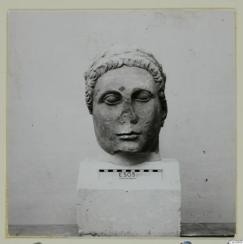 Carved stone head of a figure with curled hair, set on a rectangular block base with a scale bar