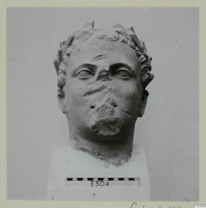 Stone head with a leafy wreath in the hair and a damaged nose and mouth, set on a white block base