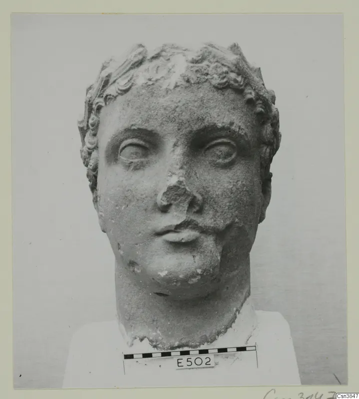 Stone head sculpture with curled hair and a raised band across the crown, shown front-on, nose broken away.