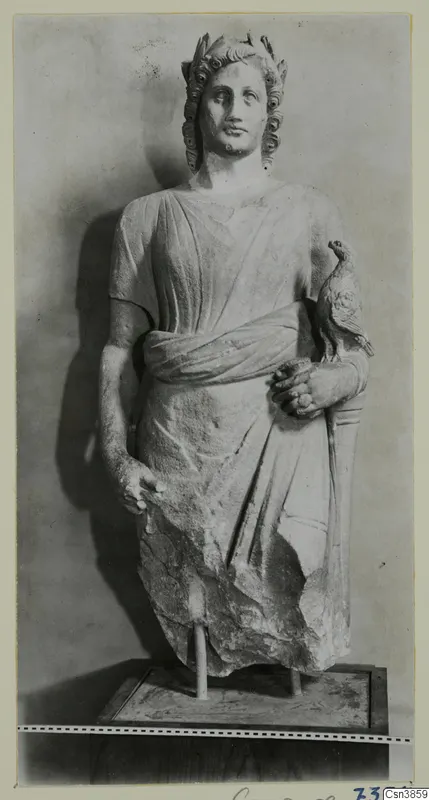 Stone statue of a draped standing figure with a bird perched on its right arm, set on a wooden base.