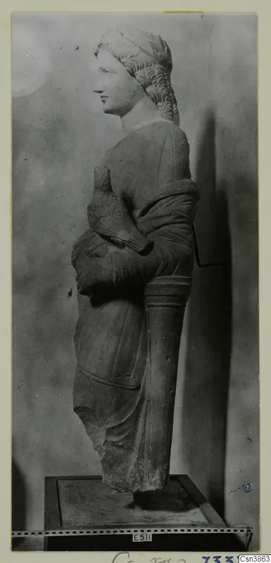 Stone statue of a female figure in left profile, with wavy hair and draped clothing, standing on a plinth.
