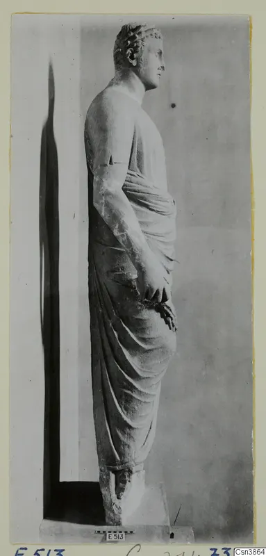 Standing draped stone statue of a figure seen in profile, facing right, on a plinth.