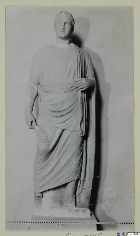 Standing stone statue of a draped figure with curled hair, photographed against a plain backdrop
