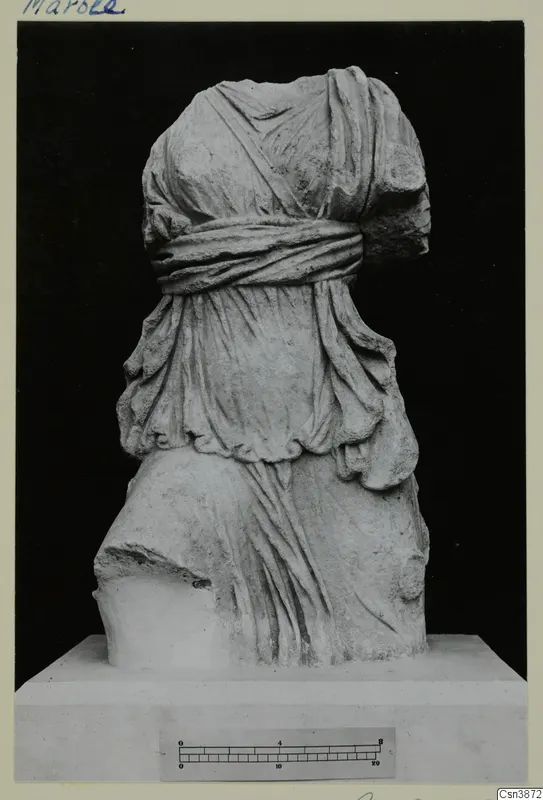 Headless draped statue in pale stone on a plinth, photographed against a black background, with a scale bar below