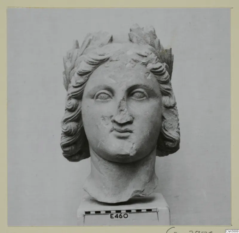Sculpted stone head with wavy hair and a leaf wreath, on a square base with a scale bar