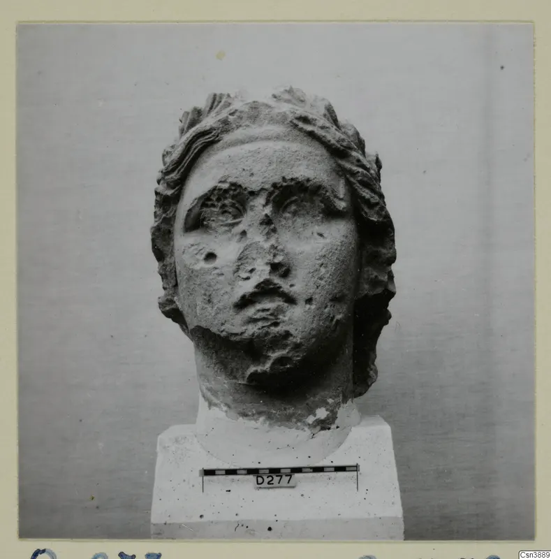 Stone head of a statue with wavy hair, shown on a plinth against a grey backdrop.