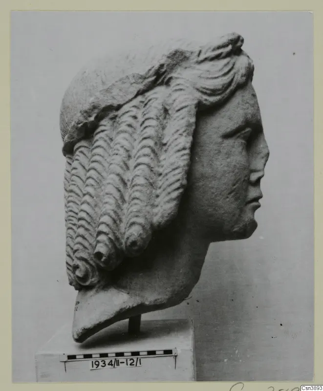 Carved stone head in profile facing right, with hair in rows of wavy curls, standing on a block base