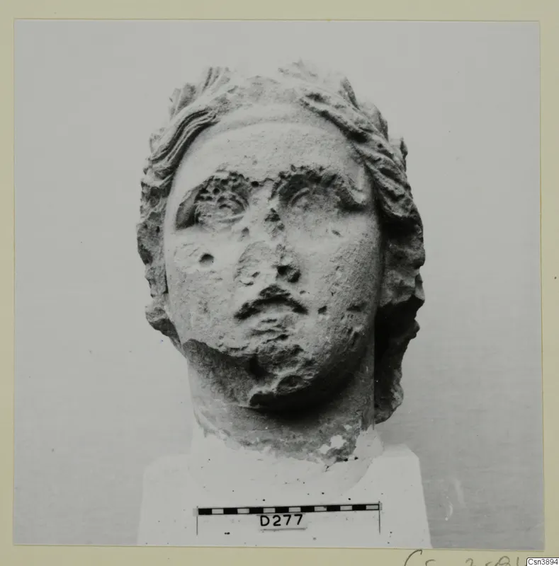 Carved stone head of a figure with wavy hair framing the face, eyes, nose and mouth worn.