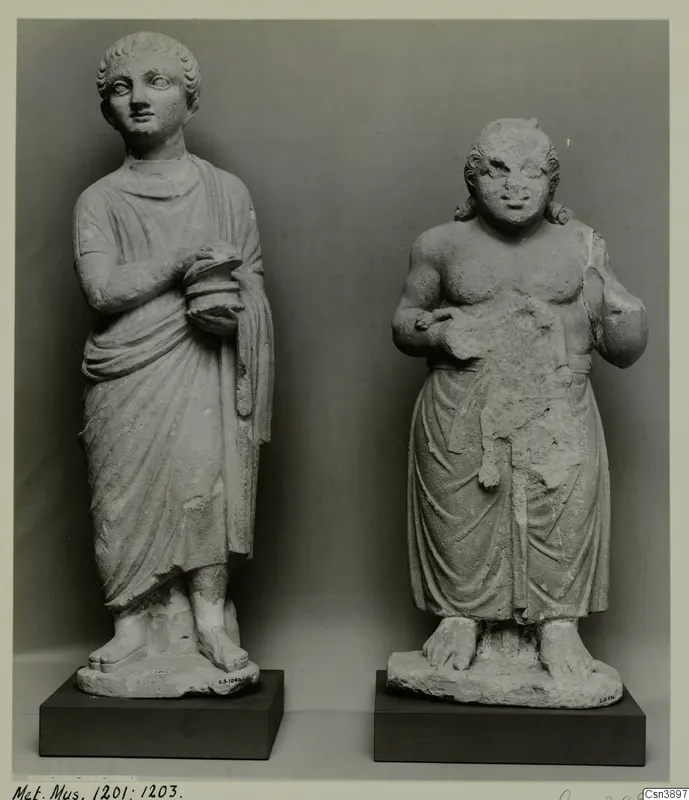 Two standing stone statues on dark bases; the left wears a draped garment and holds a round object, the right is bare-chested.