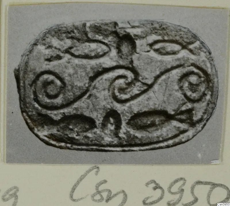 Oval seal or gem with raised spiral scrolls and curved leaf-like shapes, photographed face-on on a card mount.
