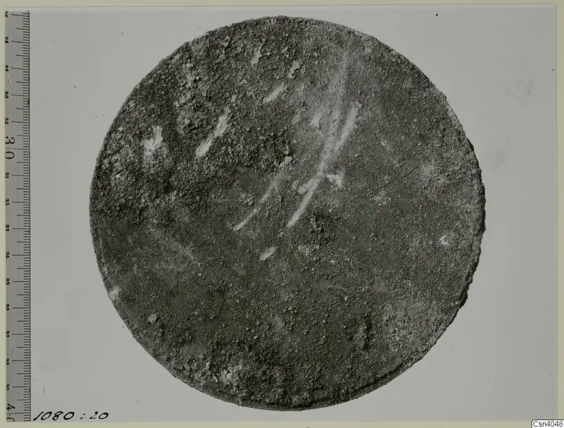 Round, corroded disc photographed beside a graduated measuring rod