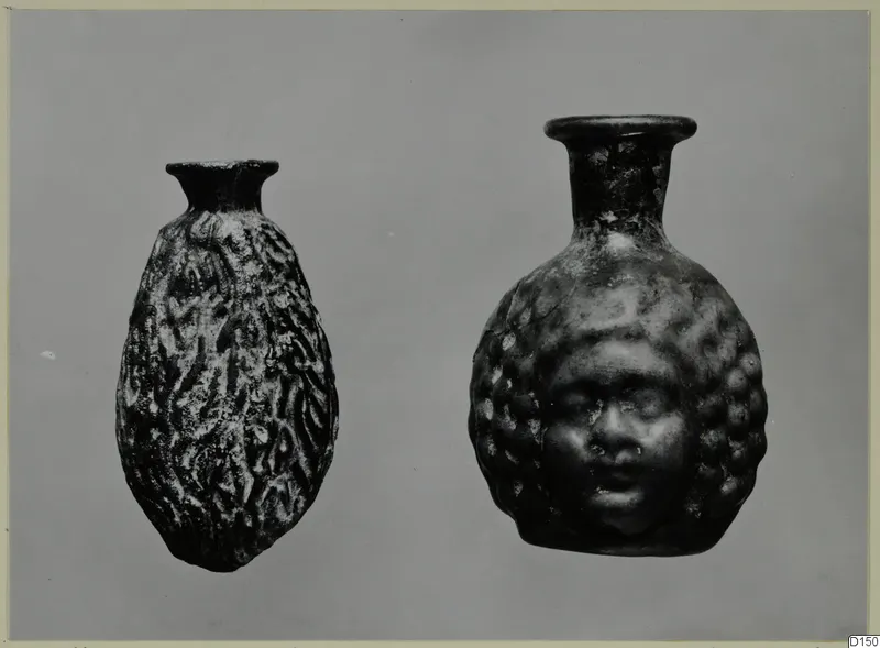 Two blown glass vessels, one with a moulded human face on its body, photographed against a grey background