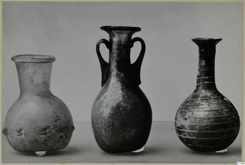 Three blown glass vessels in a row: a flared-rim bottle, a two-handled vessel and a ribbed bottle with a long neck