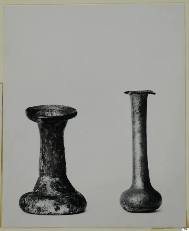 Two small glass vessels side by side on a pale background, one with a flared foot and short neck, one tall and slender-necked.