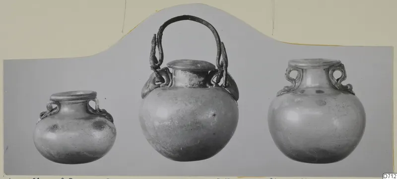 Three blown glass vessels with handles, shown side by side on a grey mount