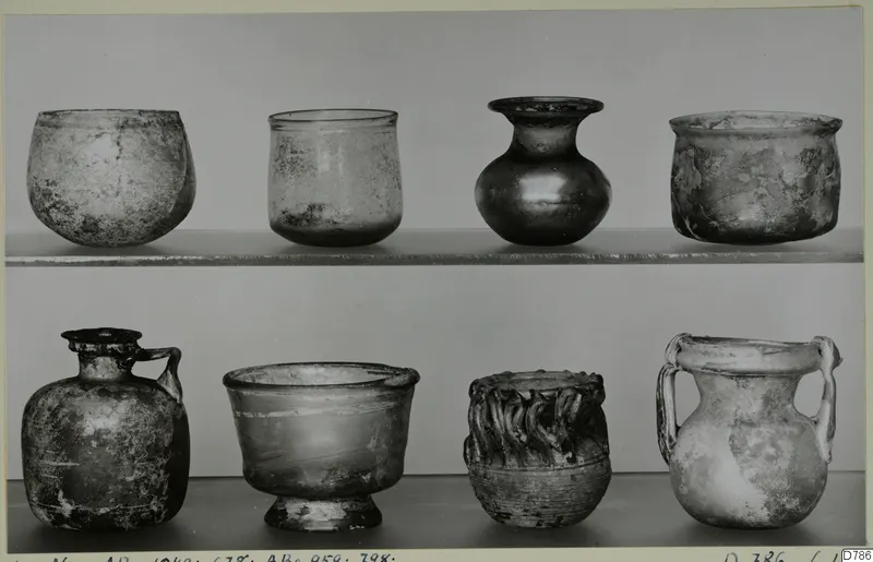 Eight blown glass vessels in two rows on shelves, including small jugs and jars with handles.