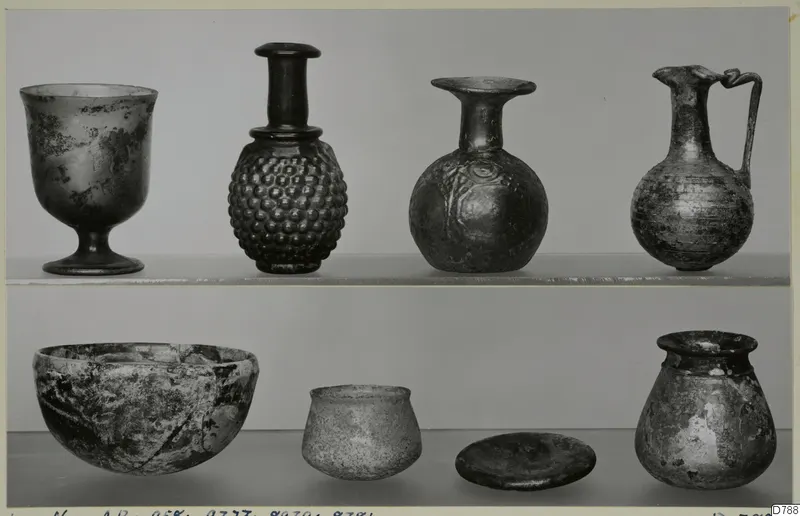 Blown glass vessels on two shelves: a footed cup, bottles, a handled jug, a deep bowl, a small cup and a jar.
